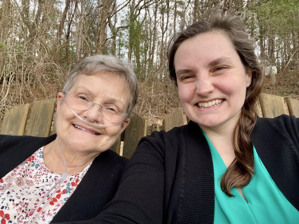 Beth, dressed in a black sweater and teal shirt, sits smiling with her mom who is wearing glasses, a black sweater, and a floral shirt.